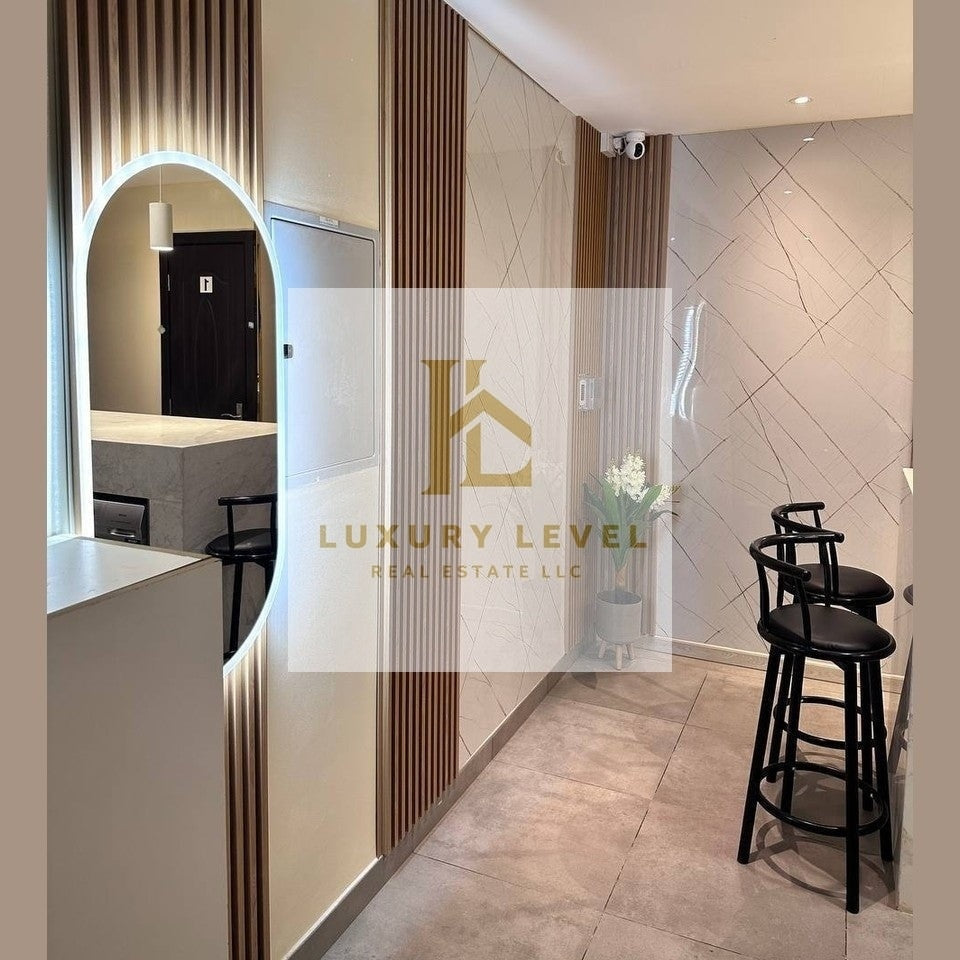 A modern interior hallway with beige stone tile floors, slatted wood wall panels, a large arched mirror with built-in lighting, and two black bar stools near a tiled wall. A semi-transparent 'LUXURY LEVEL REAL ESTATE LLC' watermark overlays the center.